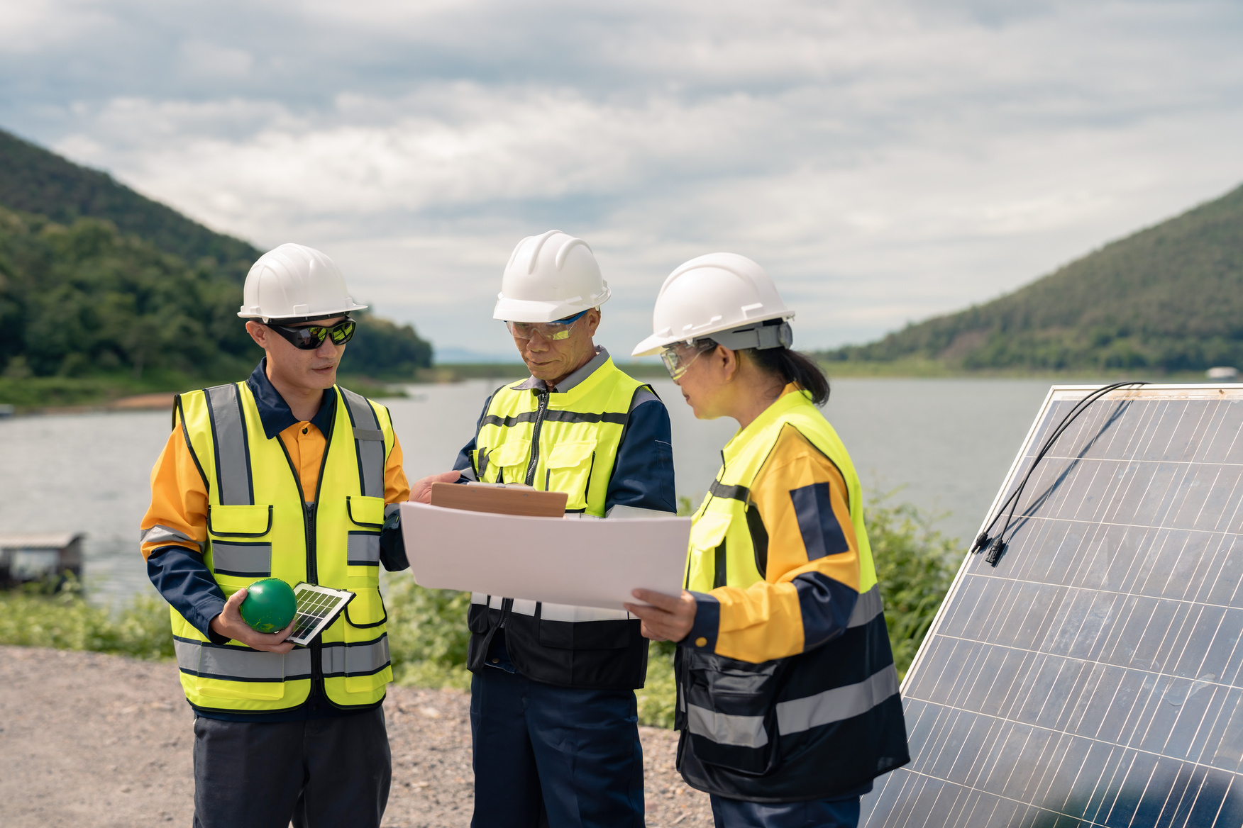 Three construction workers in hard hats and high-visibility vests examine blueprints near a solar panel alongside a body of water.